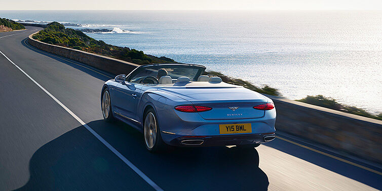 Bentley Stockholm New Bentley Continental GTC Azure convertible rear view in Light Windsor blue paint with 22 inch ten spoke wheels driving dynamically on an ocean road in the sunshine